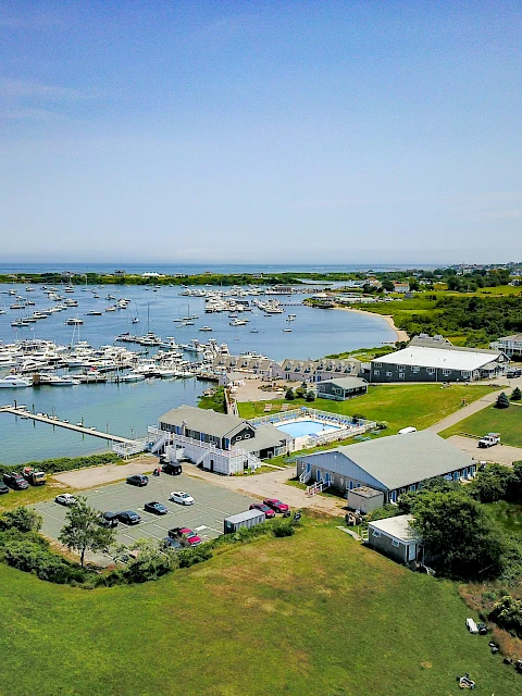 Aerial view of a marina with numerous boats, docks, and waterfront buildings; green lawns, scattered trees, and a calm coastal landscape.