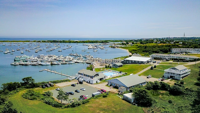 Aerial view of a marina with numerous boats, docks, and waterfront buildings; green lawns, scattered trees, and a calm coastal landscape.