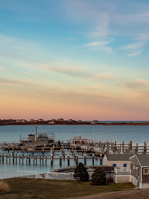 A coastal town sunset over calm harbor with boats, docks, and gray cottages along the shoreline, reflecting warm oranges and pinks.
