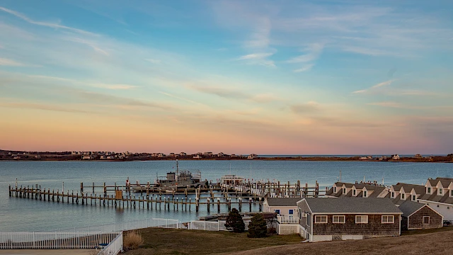 A coastal town sunset over calm harbor with boats, docks, and gray cottages along the shoreline, reflecting warm oranges and pinks.