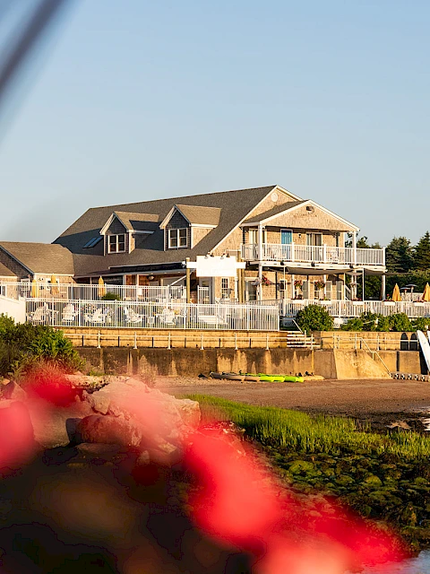 A large waterfront house with a wraparound deck sits by the water, calm waves, and a sandy shore; red flowers blur in the foreground.