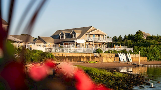 A large waterfront house with a wraparound deck sits by the water, calm waves, and a sandy shore; red flowers blur in the foreground.