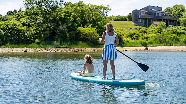 Two people stand on a paddleboard in calm water: an adult woman with a paddle and a child sitting at the front, near a green shoreline with houses in the background.