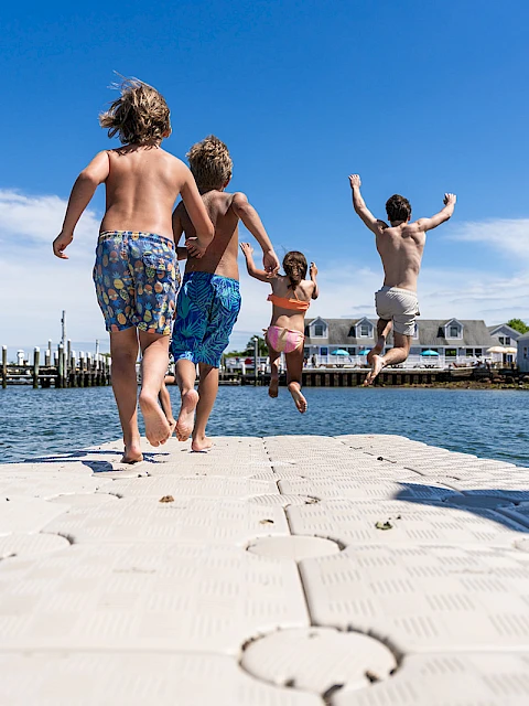 A group of kids runs barefoot on a floating dock toward the water, ready to dive and splash under a bright blue sky.