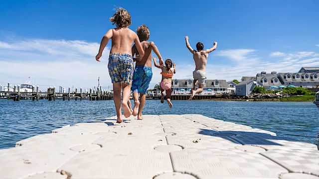 A group of kids runs barefoot on a floating dock toward the water, ready to dive and splash under a bright blue sky.