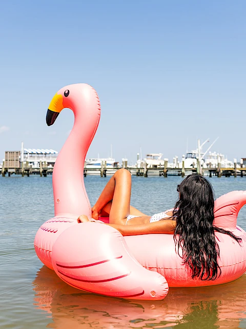 A woman lounges on a giant pink flamingo float in calm water near a marina under a clear blue sky.