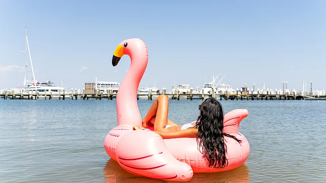 A woman lounges on a giant pink flamingo float in calm water near a marina under a clear blue sky.