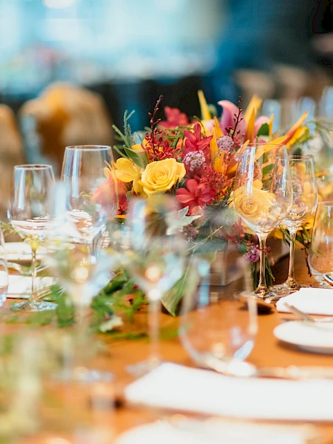 A long dining table set for a formal event with floral centerpieces, fine china, glassware, and neatly folded napkins on each place setting, all in warm lighting.