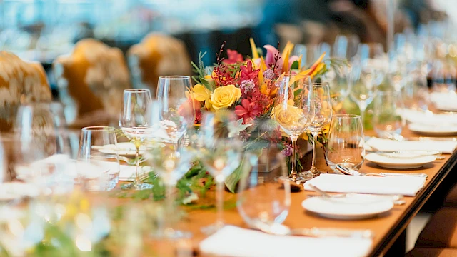A long dining table set for a formal event with floral centerpieces, fine china, glassware, and neatly folded napkins on each place setting, all in warm lighting.