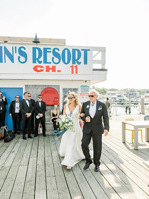 A bride in a white gown and a suited groom walk hand in hand down a wooden boardwalk, wedding guests in the background near a blue seaside resort sign, sunny day.