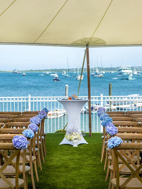 An outdoor seaside wedding setup with wooden chairs facing a blue sea, a white tall cake table lined with purple flowers under a canopy.