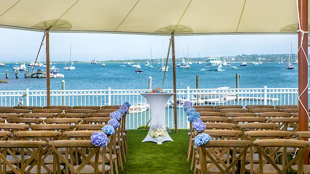 An outdoor seaside wedding setup with wooden chairs facing a blue sea, a white tall cake table lined with purple flowers under a canopy.