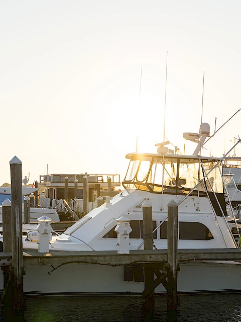 A calm marina with a white motor yacht at a dock, masts and boats in the background, and a bright sun framing the scene.