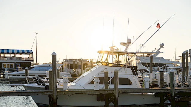 A calm marina with a white motor yacht at a dock, masts and boats in the background, and a bright sun framing the scene.
