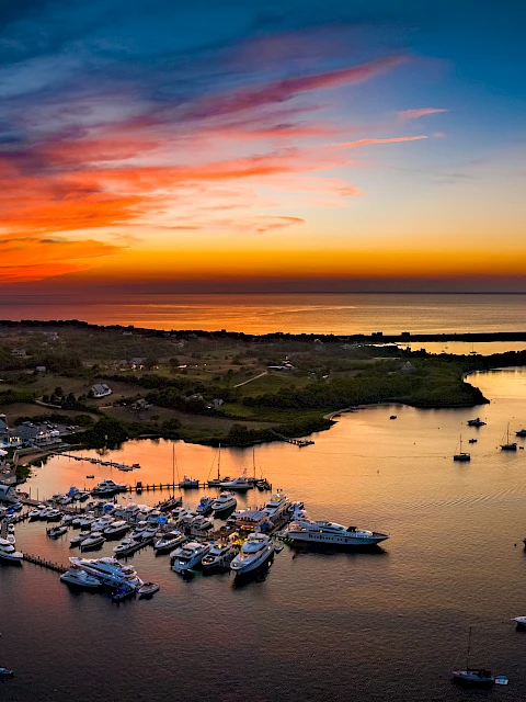 A coastal sunset with an orange-to-pink sky over calm waters, boats clustered by shore and a few rocky islets silhouetted at dusk.