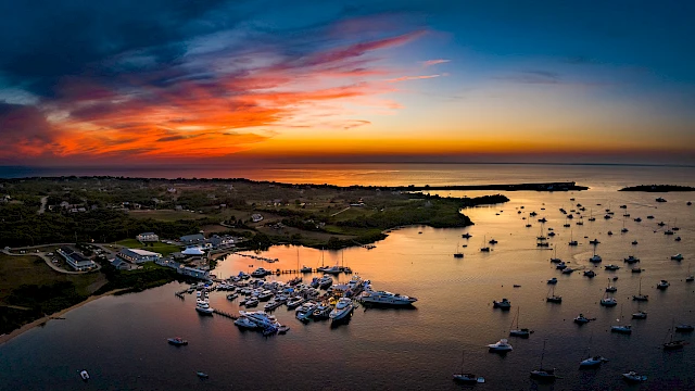 A coastal sunset with an orange-to-pink sky over calm waters, boats clustered by shore and a few rocky islets silhouetted at dusk.