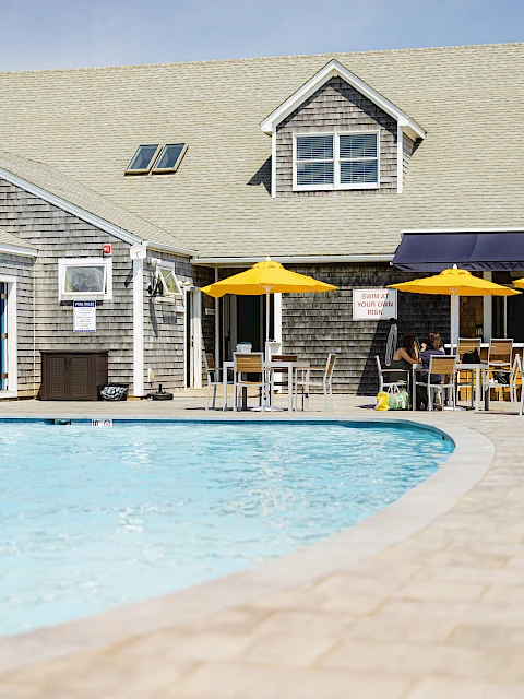 A sunny poolside scene at a clubhouse with a curved pool, wooden deck, and outdoor seating under yellow and navy umbrellas by the building.