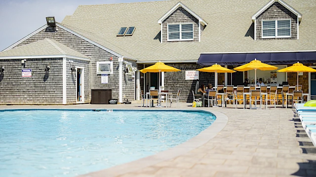 A sunny poolside scene at a clubhouse with a curved pool, wooden deck, and outdoor seating under yellow and navy umbrellas by the building.