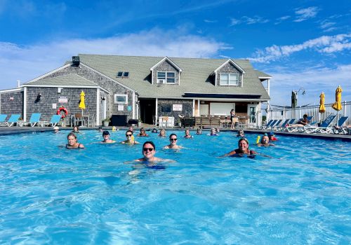People swim and relax in a large outdoor pool in front of a gray two-story building with yellow umbrellas and lounge chairs, sunny day.