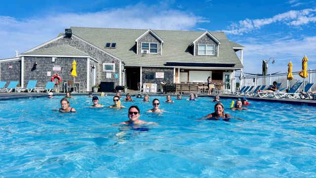People swim and relax in a large outdoor pool in front of a gray two-story building with yellow umbrellas and lounge chairs, sunny day.