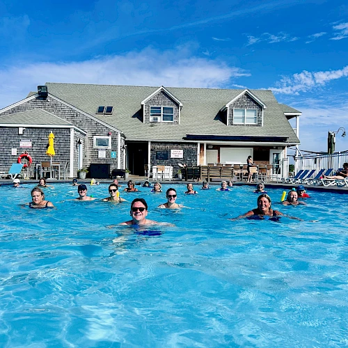 People swim and relax in a large outdoor pool in front of a gray two-story building with yellow umbrellas and lounge chairs, sunny day.