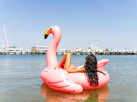 A woman lounges on a pink inflatable flamingo float in calm water beside a marina.