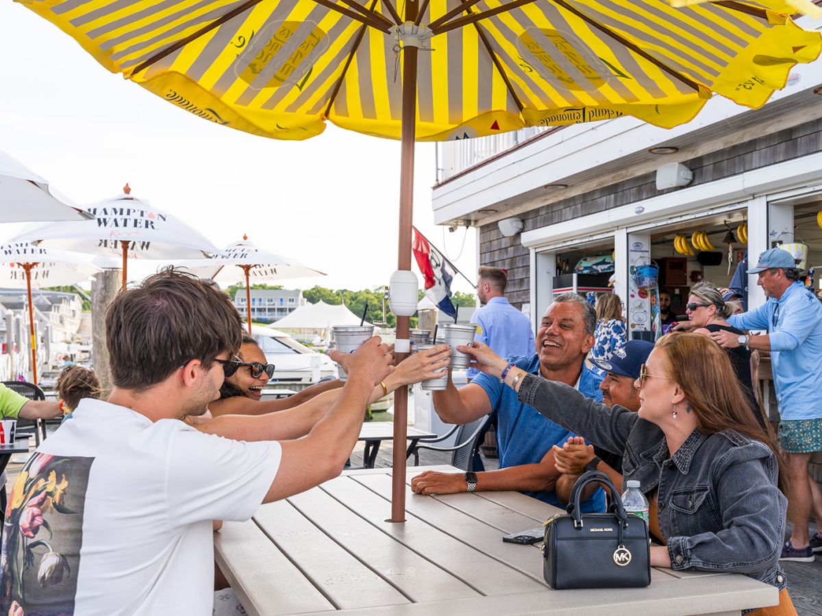 Friends cheering drinks at an outdoor bar, with yellow striped umbrellas overhead in sunny weather, at a lively waterfront setting.