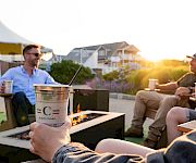 Two friends lounge by a backyard fire pit, sipping from cups with straws at sunset.