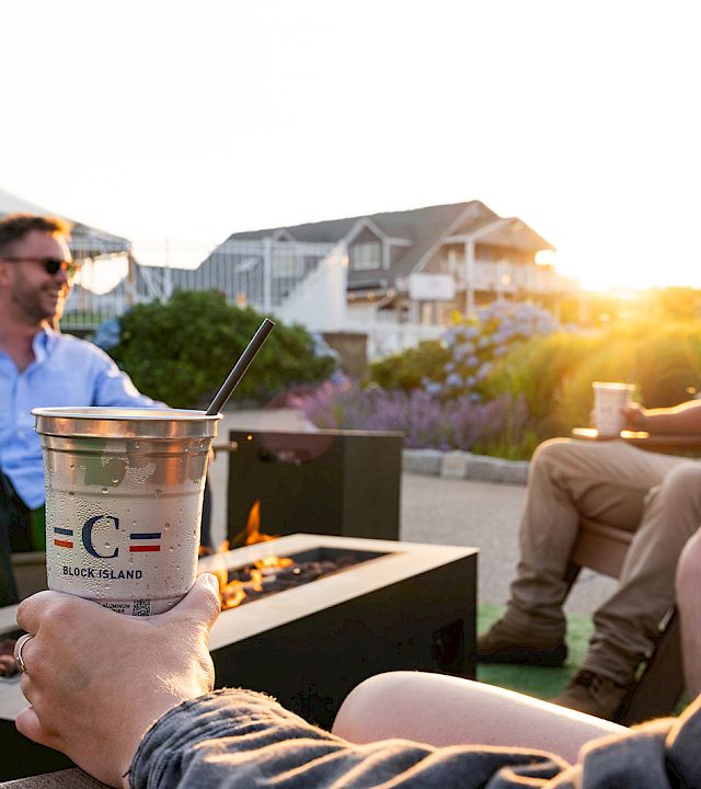 Two friends lounge by a backyard fire pit, sipping from cups with straws at sunset.