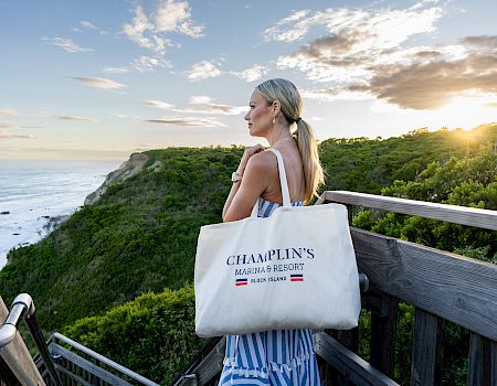 Woman in striped dress on a seaside overlook at sunset, holding a tote bag labeled “Champlin’s Marina & Resort”.