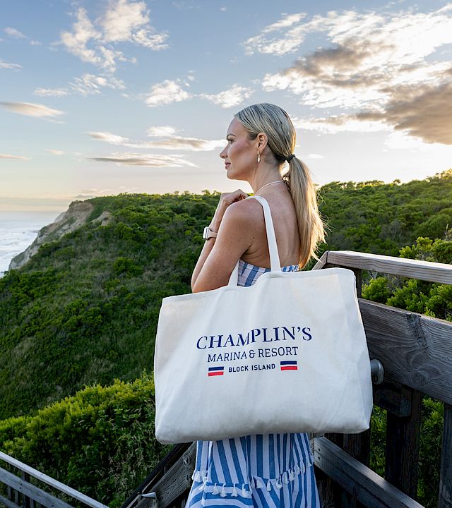 Woman in striped dress on a seaside overlook at sunset, holding a tote bag labeled “Champlin’s Marina & Resort”.
