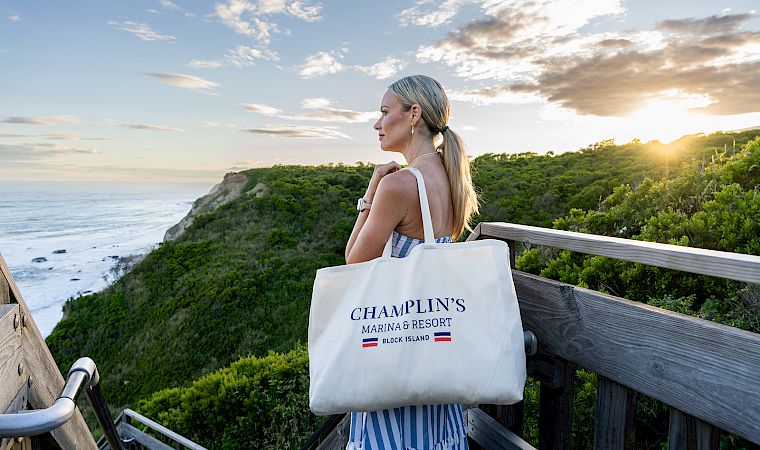 Woman in striped dress on a seaside overlook at sunset, holding a tote bag labeled “Champlin’s Marina & Resort”.