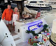 Two people pose on a holiday-decorated yacht at a marina, with an American flag and twinkling lights on the boat.