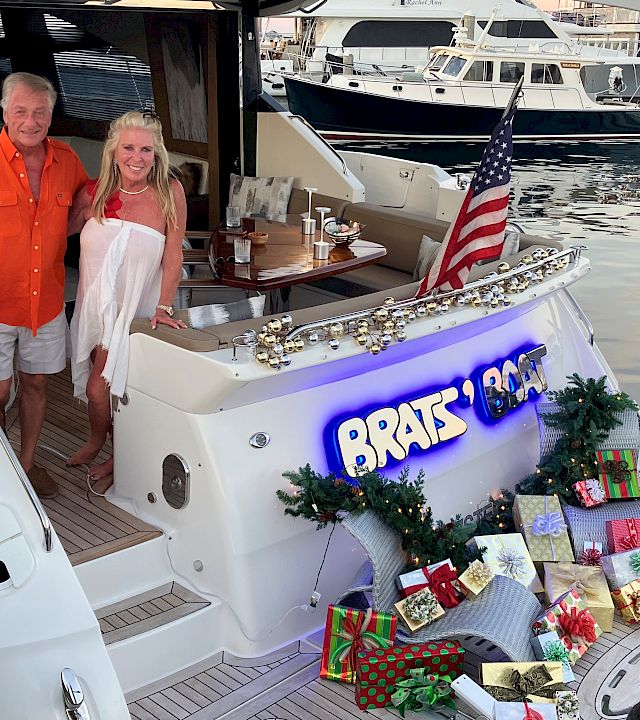 Two people pose on a holiday-decorated yacht at a marina, with an American flag and twinkling lights on the boat.