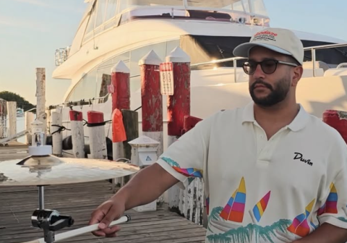 A man in sunglasses plays steel drums on a sunny dock beside a large boat, with colorful posts in the background.