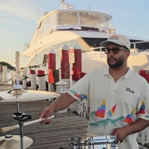 A man in sunglasses plays steel drums on a sunny dock beside a large boat, with colorful posts in the background.