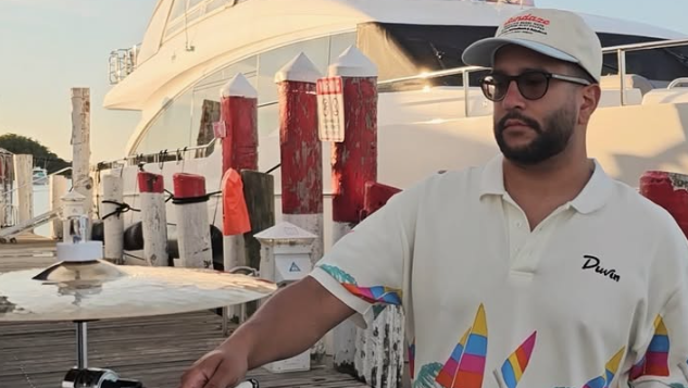 A man in sunglasses plays steel drums on a sunny dock beside a large boat, with colorful posts in the background.