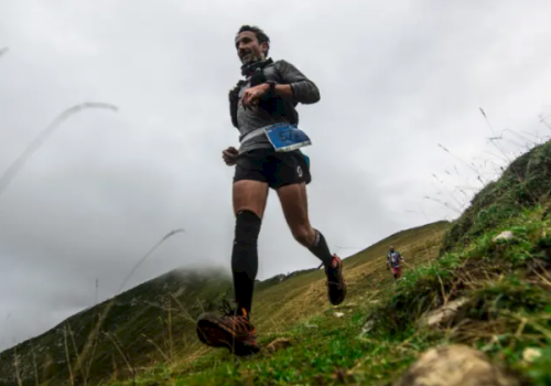 A runner sprints uphill on a grassy hill, arms pumping as cloudy skies loom behind him.
