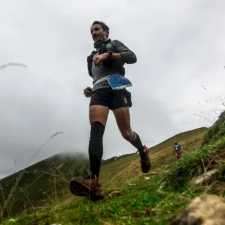 A runner sprints uphill on a grassy hill, arms pumping as cloudy skies loom behind him.
