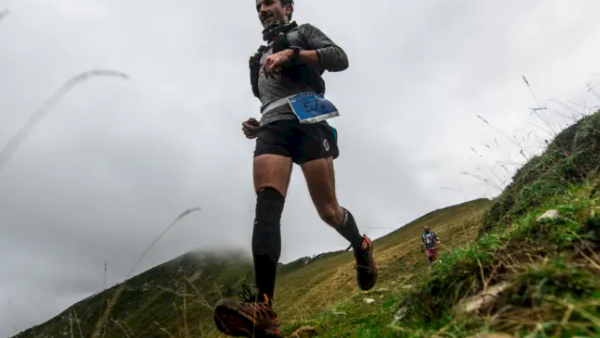 A runner sprints uphill on a grassy hill, arms pumping as cloudy skies loom behind him.