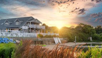 A house near a lakeside with a beautiful sunset in the background, surrounded by greenery and flowers, creating a serene atmosphere.