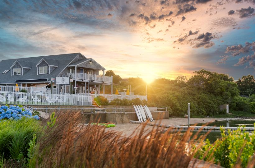 A house near a lakeside with a beautiful sunset in the background, surrounded by greenery and flowers, creating a serene atmosphere.