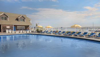 The image depicts a poolside area with lounge chairs, yellow umbrellas, a house-like building, and a waterfront view under a clear blue sky.